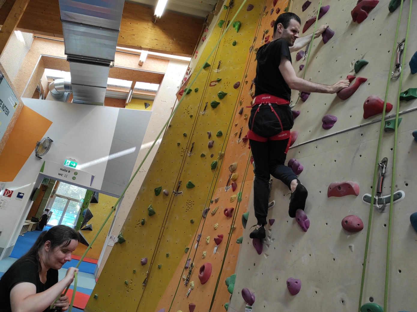 Ein Mann auf einer indoor Kletterwand wird von einer Frau mit einem Seil und Klettergurten gesichert. Die Kletterwand hat viel bunte Griffe. Mehrere Kletterseile mit Karabinern an den Enden hängen von der Kletterwand. Im Hintergrund ist ein Durchgang ins Freie und eine Lüftungsanlage an der Hallendecke sichtbar.