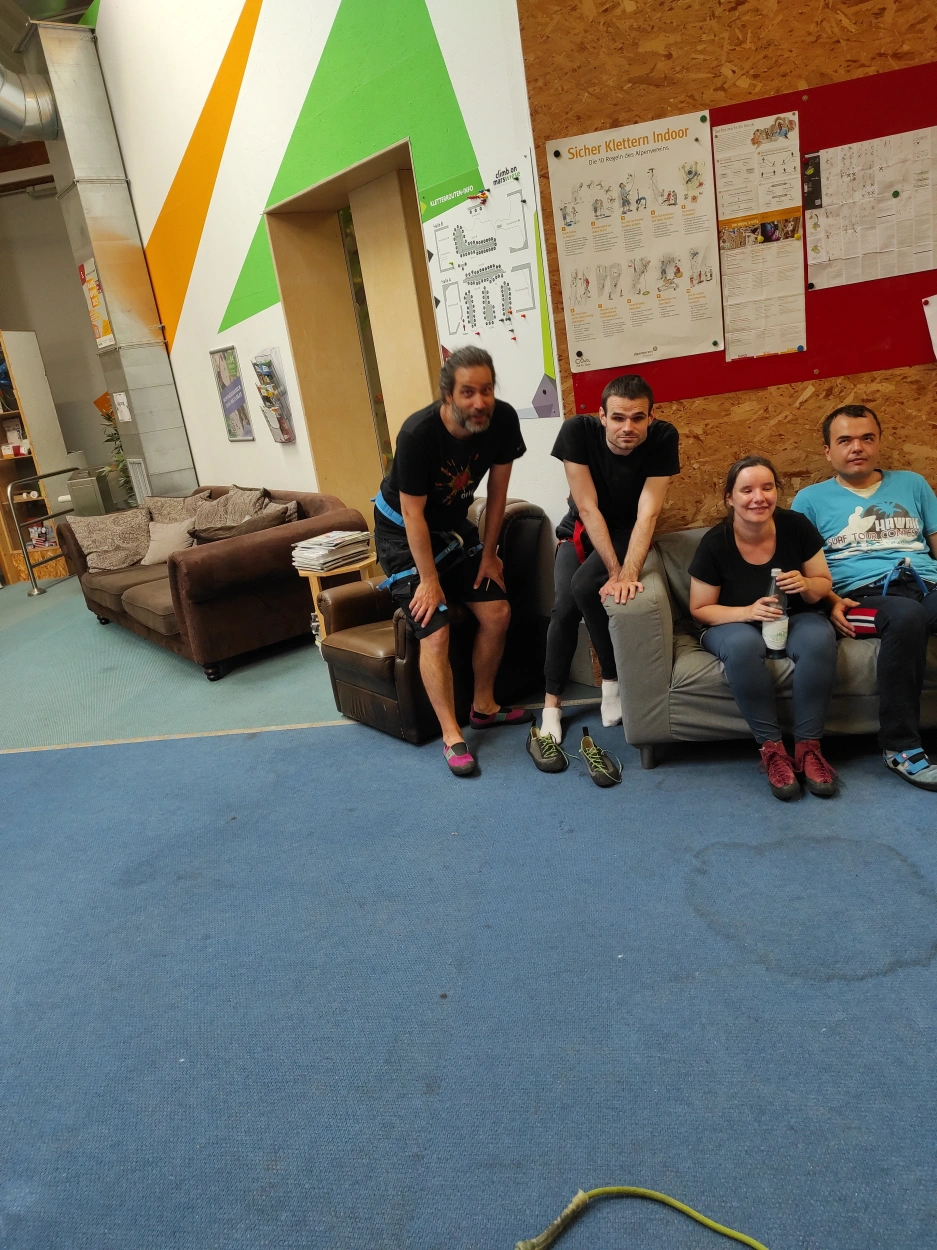 Gruppenfoto von vier Personen mit Kletterschuhen und Klettergurten auf einer Sitzgruppe. Die Frau und die drei Männer lächeln in die Kamera und wirken etwas erschöpft. Die Wand dahinter besteht teilweise aus Grobspanplatten mit einer Pinnwand und zum anderen Teil aus einer weißen Wand mit aufgemalten einfachen geometrischen Flächen. Den Boden bildet ein blauer Teppich.