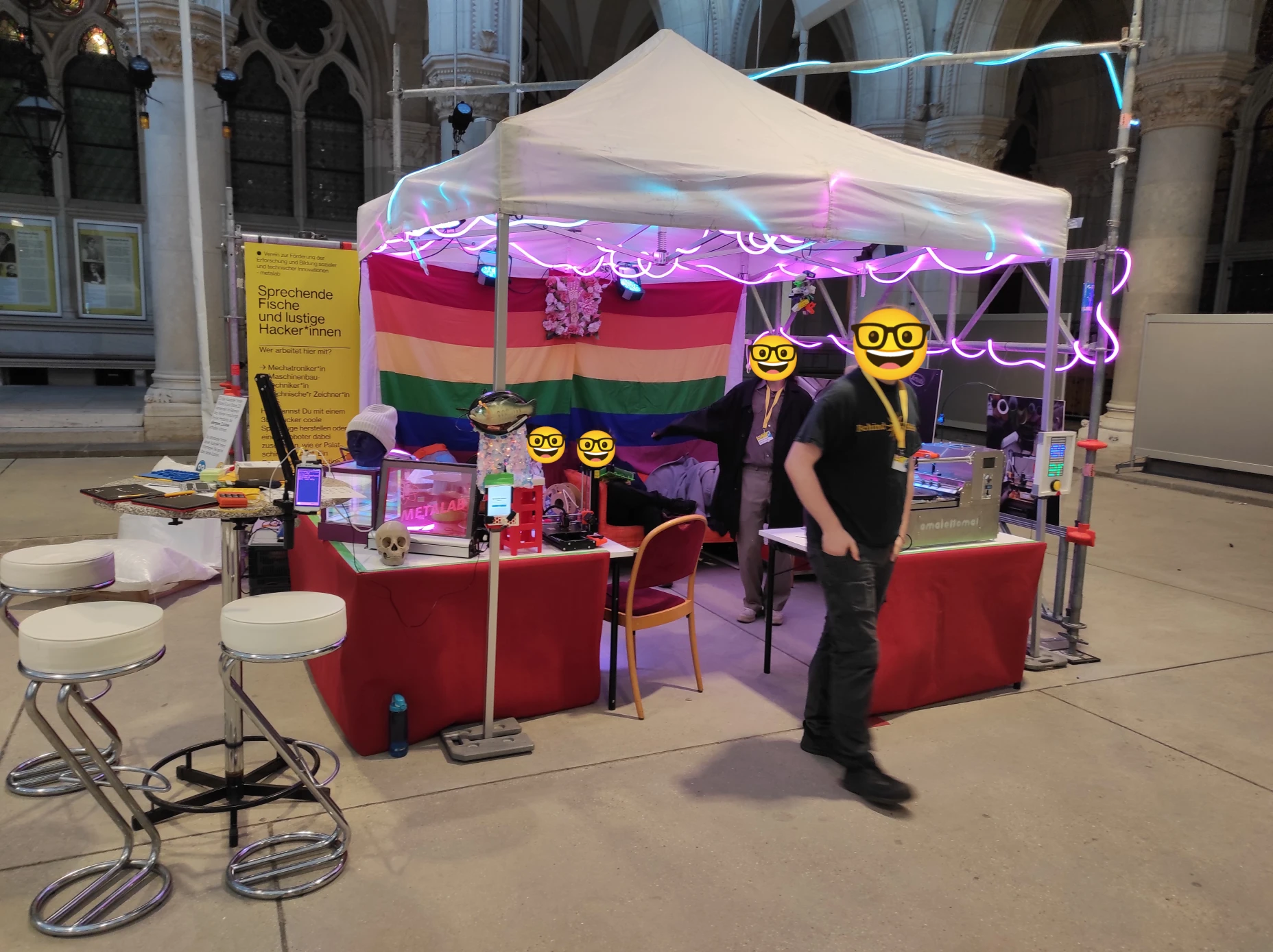Vier mit Nerdsmiley anonymisierte Personen an einem Stand mit Zeltdach. Eine Regenbogenfahne mit einem Relief aus Blumen und gebärdenden Händen in der Mitte bildet die Standrückwand. Rosa und hellblau leuchtende Lichtschlangen sowie zwei Scheinwerfer hängen im Zeltdach und im umrahmenden Baustahlgerüst. Im Hintergrund befinden sich gotische Fenster und Säulen aus Sandstein. Auf den mit rotem Stoff eingekleideten Tischen steht eine Apparatur mit Metallgehäuse und der Aufschrift amalettomat sowie ein 3D-Drucker, eine Telefonzelle aus Kunststoff, ein Leuchtschild mit dem Logo vom metalab, ein Totenkopf, ein Kopf mit Mütze, und ein halbtransparenter Würfel der von innen leuchtet. Am Zeltgestänge ist ein sprechender Fisch und ein Display mit großem Druckknopf befestigt. Auf einem gelben Schild steht als Überschrift „Sprechende Fische und lustige Hacker*innen“. Auf einem Stehtisch mit Barhockern vor dem Stand liegen Tafeln, eine Brailletastatur und ein Smartphone ist auf einem verstellbarem Arm am Tisch befestigt.