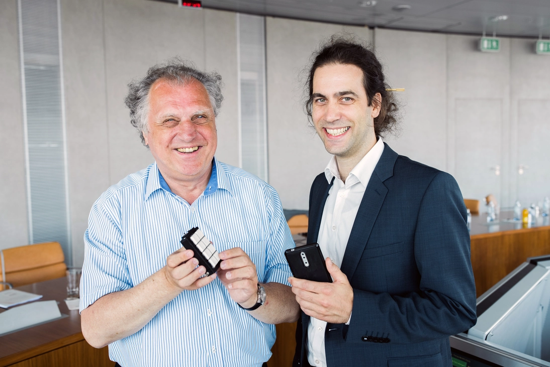 Zwei Männer lächeln strahlend in die Kamera. Sie stehen in einem Konferenzraum mit grauen Wänden und braunen Tischen. Der Mann links, Erich Schmid, im Bild ist älter, hat graue Haare und ein weiß, blau gestreiftes Hemd an. Er präsentiert ein schwarzes Kästchen mit acht weißen Tasten mit beiden Händen. Der Mann rechts im Bild hat hochgesteckte, dunkle Haar, trägt ein weißes Hemd und ein Sakko. Er hält ein Smartphone in das Bild.