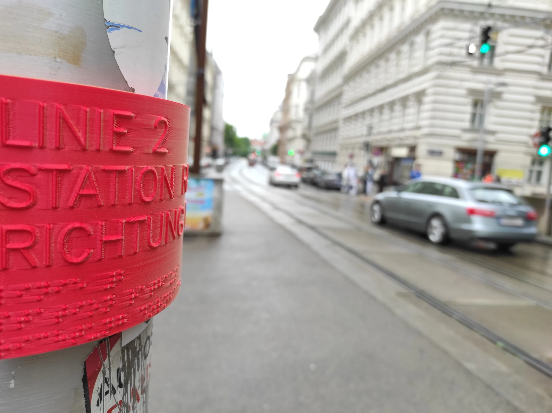 Auf einem 3D-gedruckten, roten Schild steht „Linie 2, Station, Richtung“ in taktiler Profilschrift und in Braille. Es ist nur ein erster Teil des Schildes erkennbar, weil das Schild rund ist und der weitere Text verdeckt ist. Das Schild befindet sich auf einem runden Mast aus Metall mit Aufklebern. Im unscharfen Hintergrund sind Verkehrsteilnehmer und grüne Ampeln in einer Straße mit Straßenbahnschienen und mehrstöckige Häuser aus der Gründerzeit erkennbar.