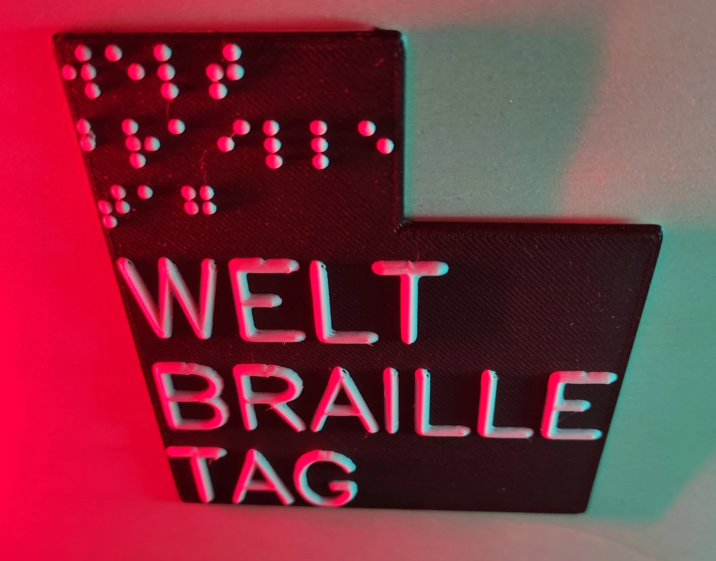 Schild aus dem 3D-Drucker „welt braille tag“ in weißer Braille- und Schwarzschrift auf schwarzem Grund. Das Schild wird von den Seiten in Rot und Blau beleuchtet. Das Schild liegt auf beigem Hintergrund.